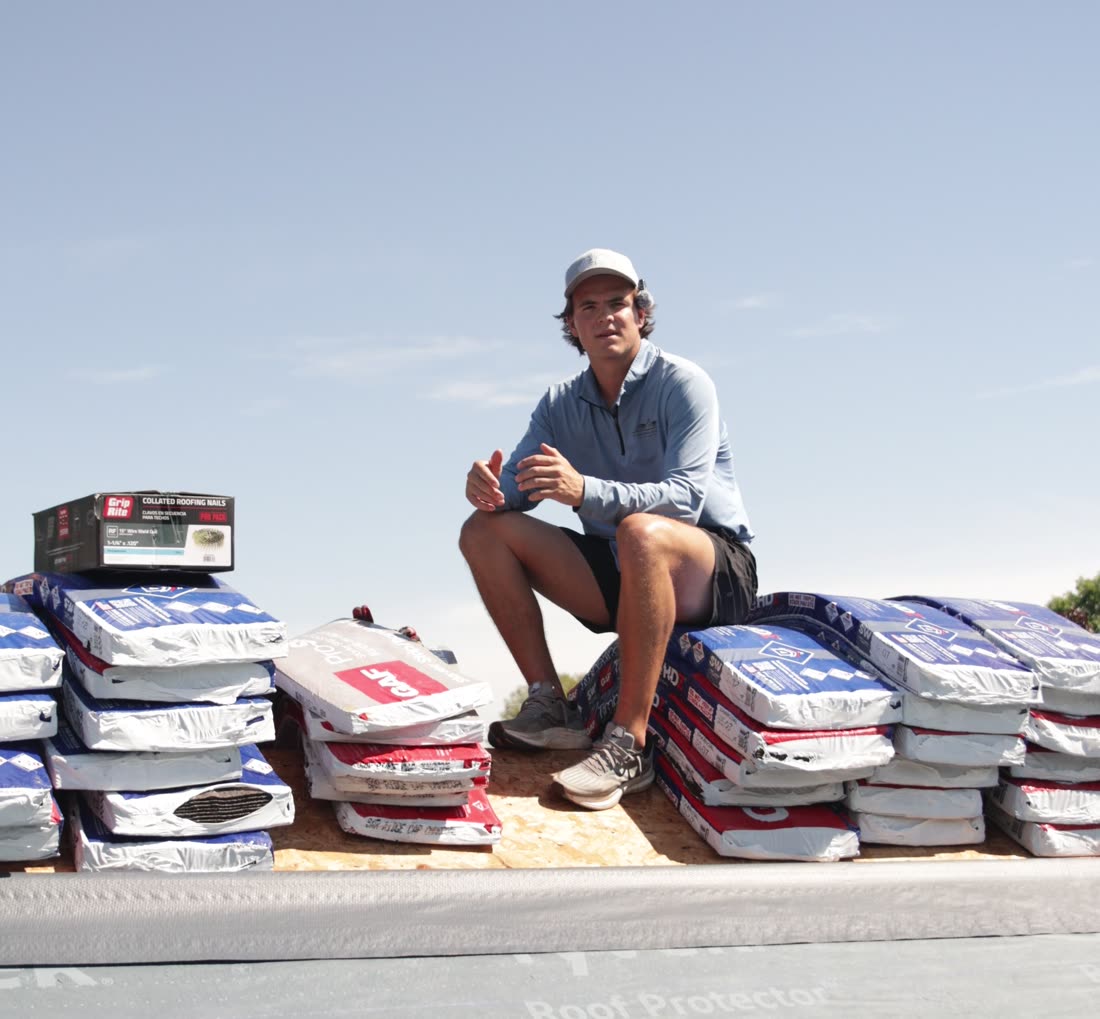 A Stoneridge team member sitting on bundles of shingles on top of a roof
